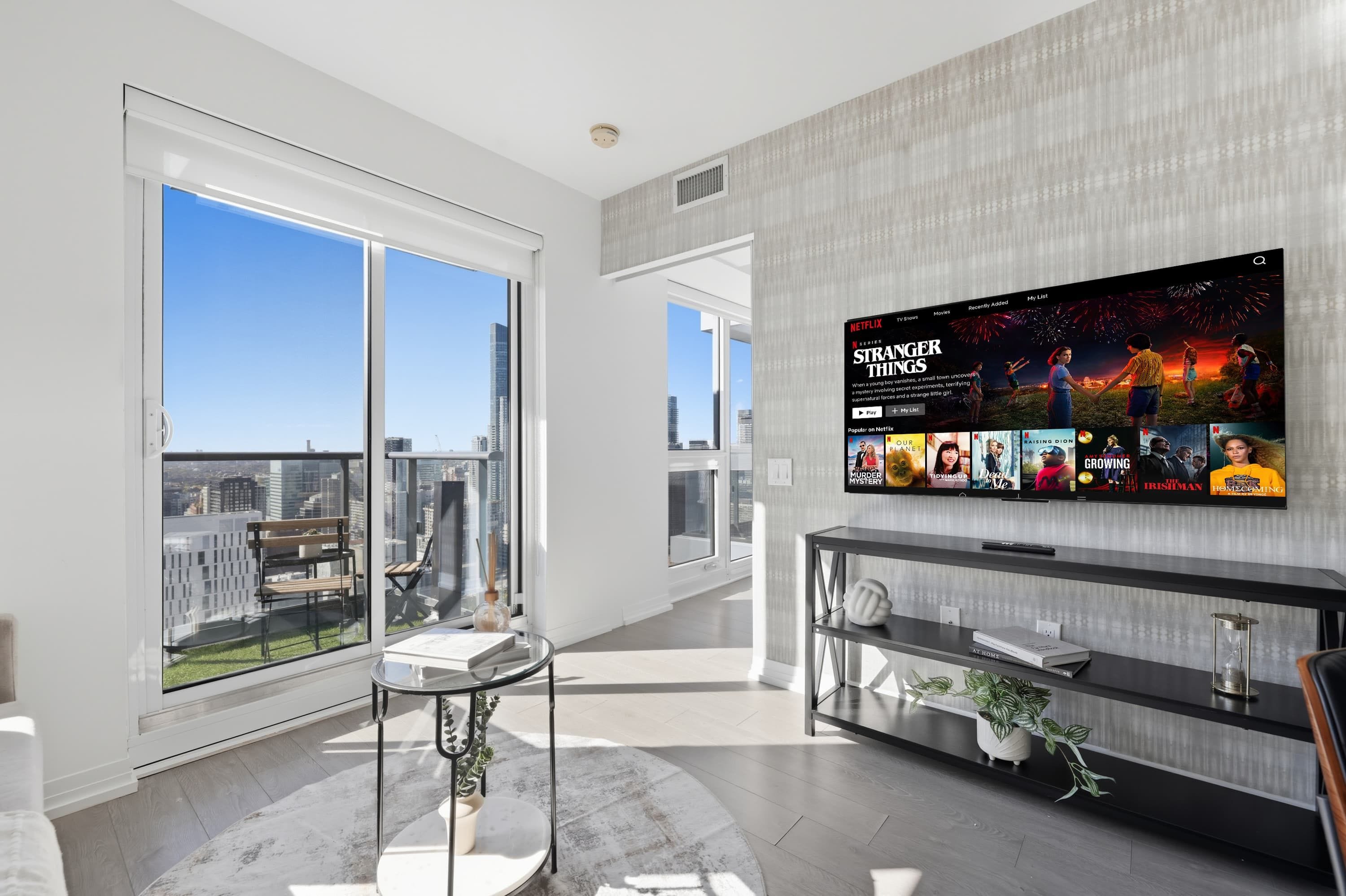 A modern living room with a large TV displaying a Netflix series, a balcony with a city view, and a minimalist decor style.