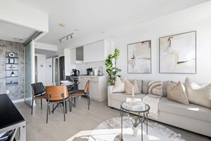 Un salon moderne avec un canapé, une table basse et des fauteuils. Il y a également un coin cuisine avec des placards et des appareils électroménagers. La pièce présente un design minimaliste, avec des murs blancs et un mur d’accent en béton.
