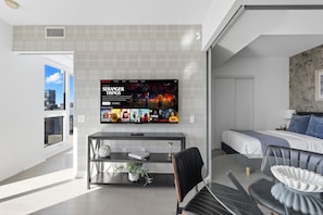 A modern hotel room with a large flat-screen TV mounted on the wall, a bed with a headboard, a bedside table with a lamp, a chair, and a potted plant. - Stunning Family-Friendly Parking & City Views! | Parking Available (Toronto)