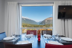 A dining table set for four with a mountain view through a large window, a flat-screen TV mounted on the wall, and a red decorative item on the table.