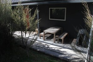 A wooden deck with a table and benches, surrounded by trees and a dark building facade.