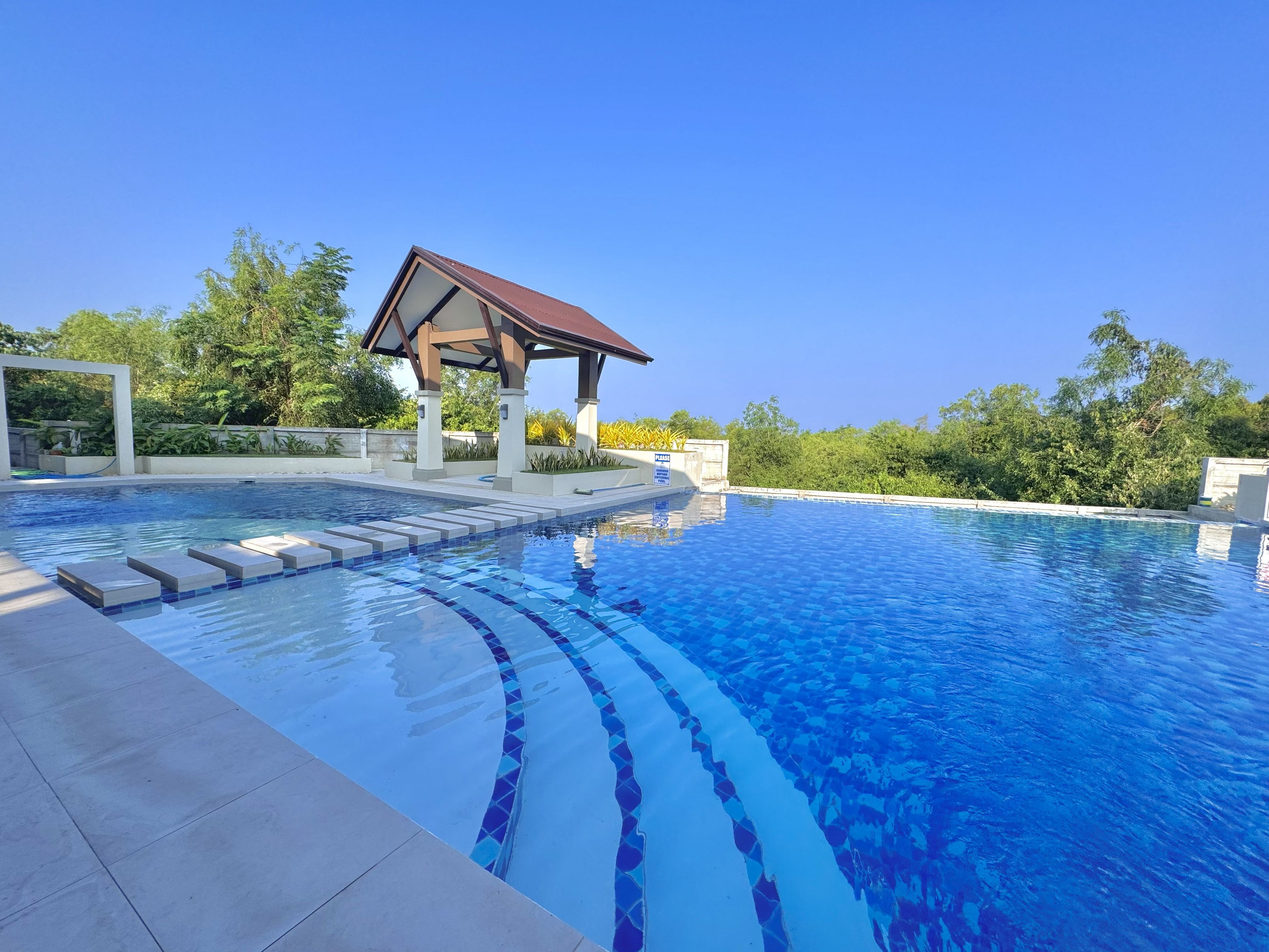 A swimming pool with a tiled edge, a covered pavilion, and surrounding greenery.