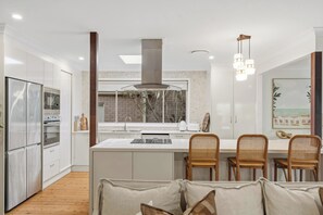 A modern kitchen with white cabinetry, a central island, and a dining area with wicker chairs. - Lakeview@macs by Coast Hosting (MacMasters Beach)