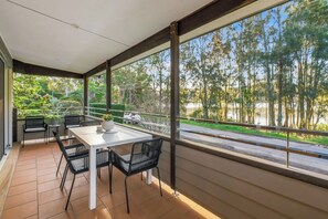 A covered outdoor dining area with a table and chairs, large windows with a view of trees and a road. - Lakeview@macs by Coast Hosting (MacMasters Beach)