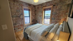 Una camera da letto con parete in mattoni, un letto con molti cuscini, una finestra e una ventola a soffitto.