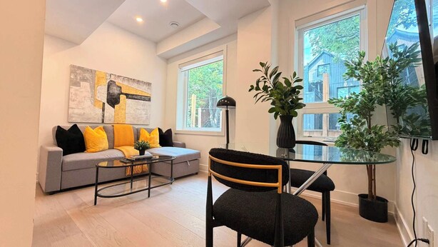Un salon moderne avec un canapé gris, des coussins noirs et jaunes, une table basse en verre, un fauteuil noir et une grande fenêtre donnant sur un immeuble.