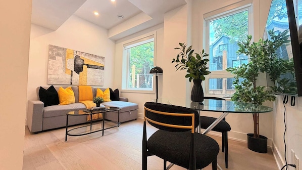 A modern living room with a grey sofa, black and yellow cushions, a glass coffee table, a black chair, and a large window with a view of a building.