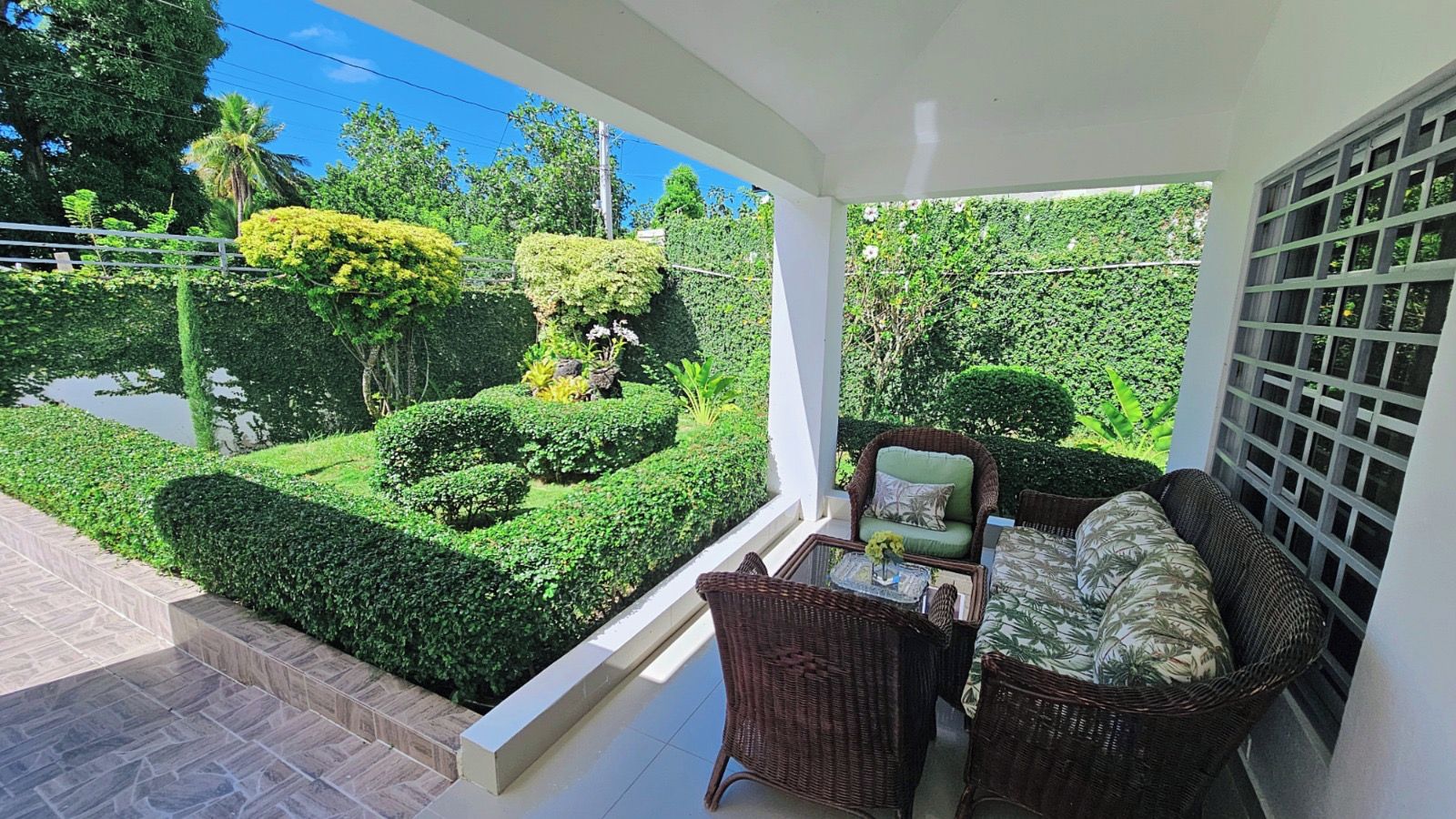 A balcony with wicker furniture, a glass table, and a view of a well-manicured garden.