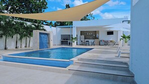 A swimming pool with a built-in waterfall feature, a sun terrace with outdoor seating, and a kitchen area visible through an open door.