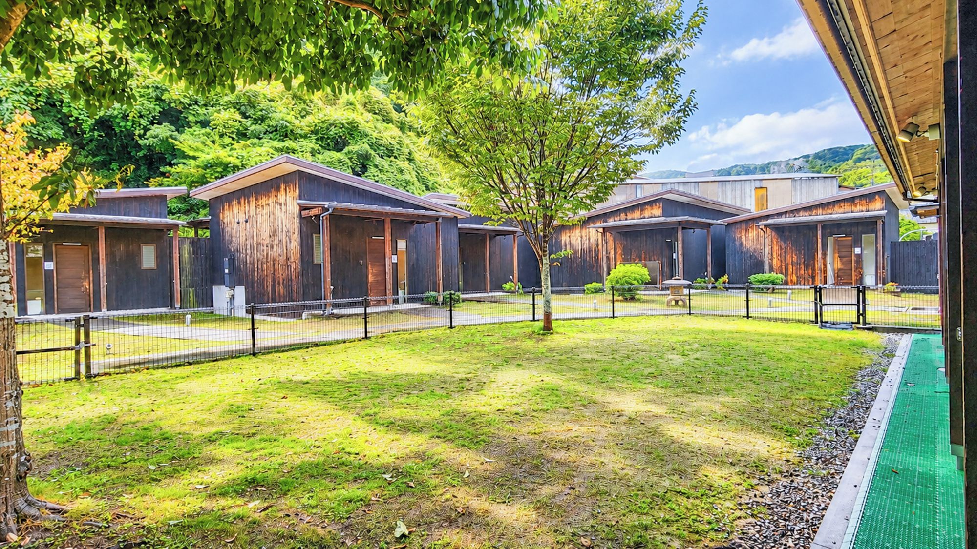 Une rangée de cabanes en bois, avec une cour verdoyante, entourée d’arbres et d’une clôture.