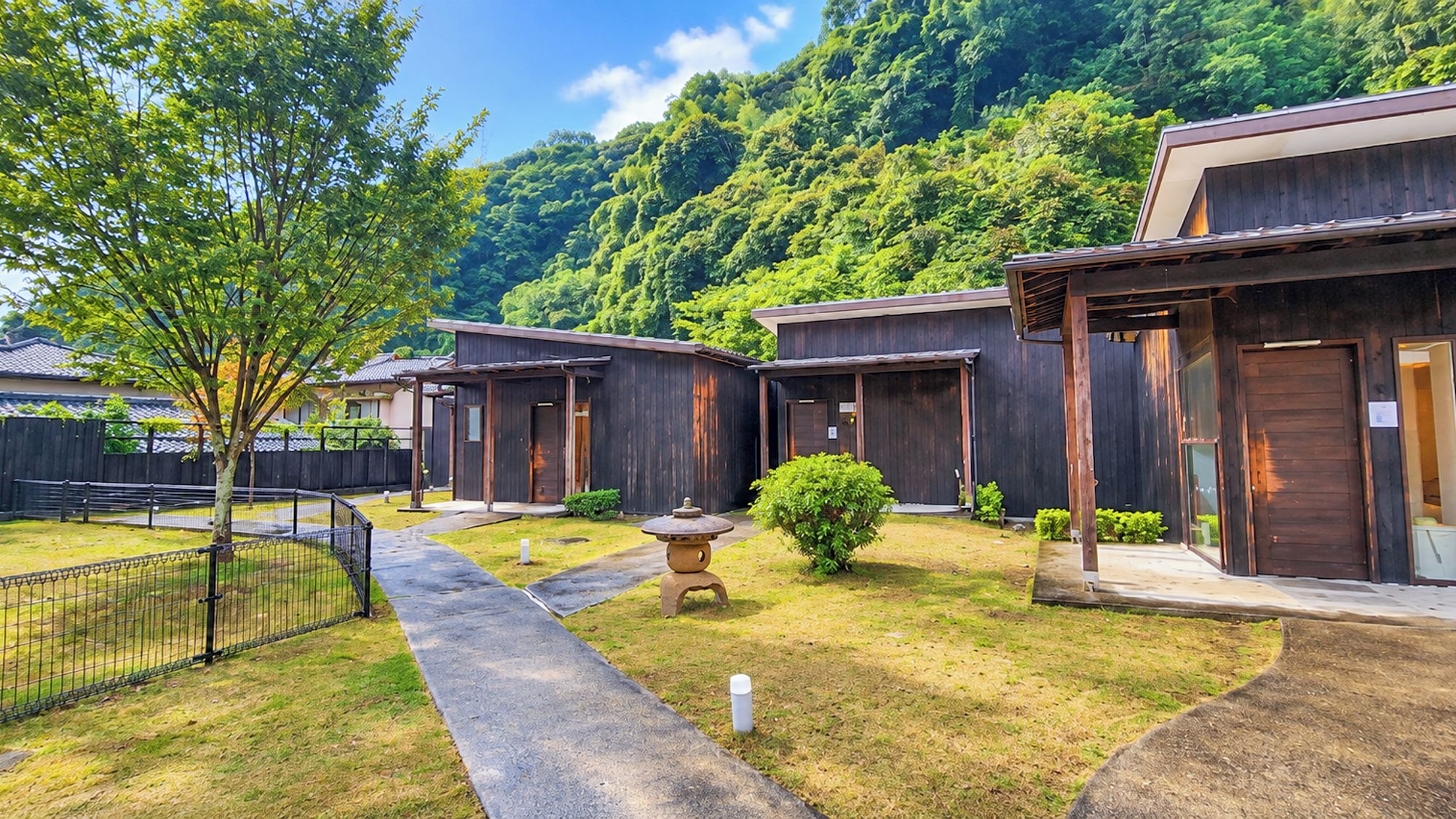 Une rangée de bâtiments en bois, avec un chemin menant à une porte, le tout entouré de verdure et bordé d’une colline boisée.