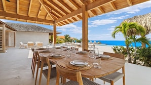 An outdoor dining area with a wooden table set for six, overlooking a beach with palm trees and the ocean. - Kas Amare (Kralendijk)