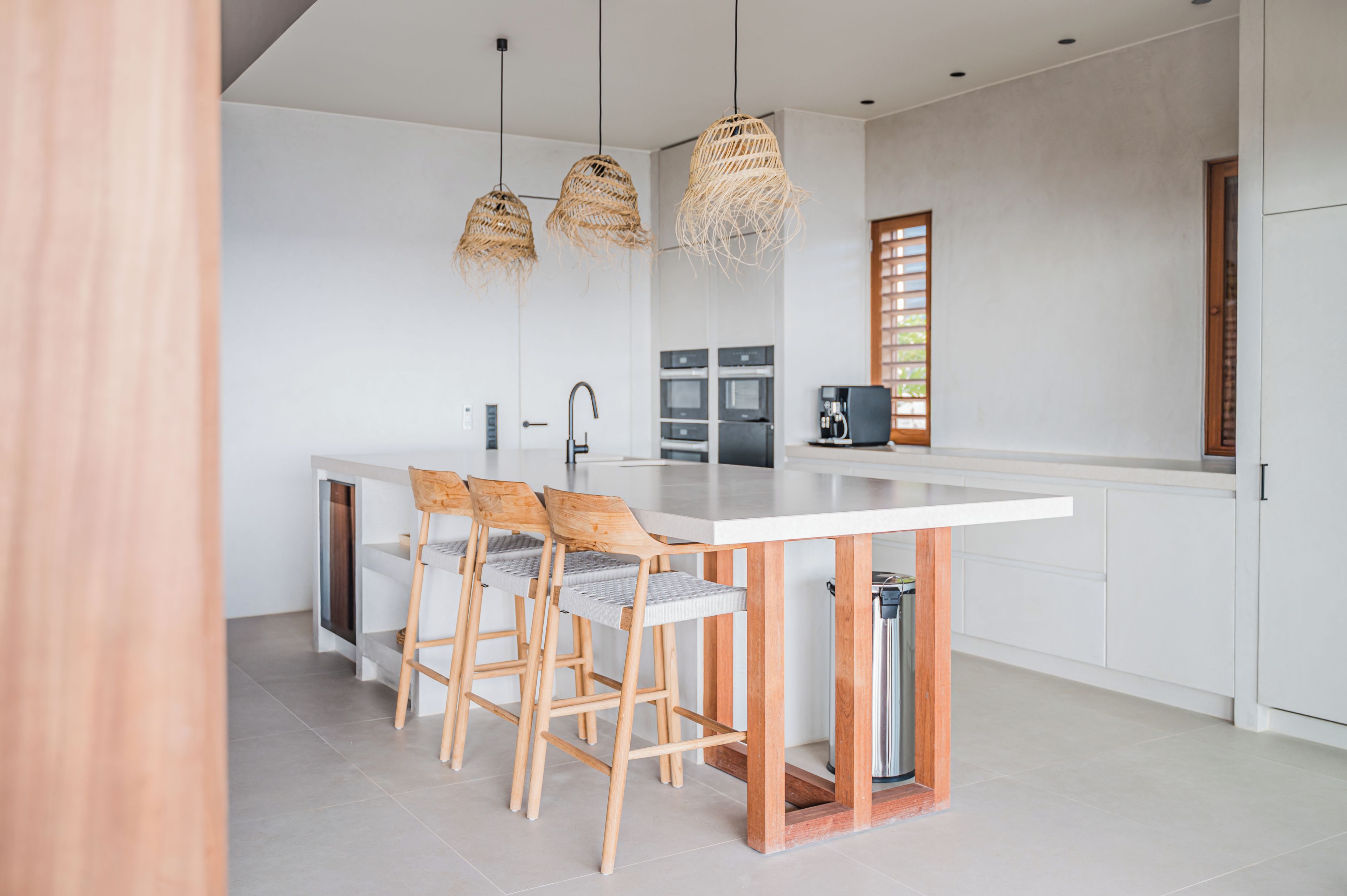 A modern kitchen with a central island, wooden chairs, and hanging wicker lights.
