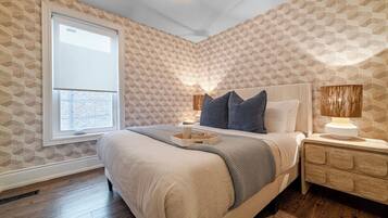 A bedroom with a large bed, a bedside table with a lamp, a window with blinds, and patterned wallpaper.