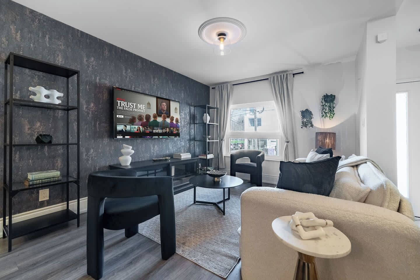 A modern living room with a dark wall, a black sofa, a coffee table, a bookshelf, and a TV mounted on the wall.