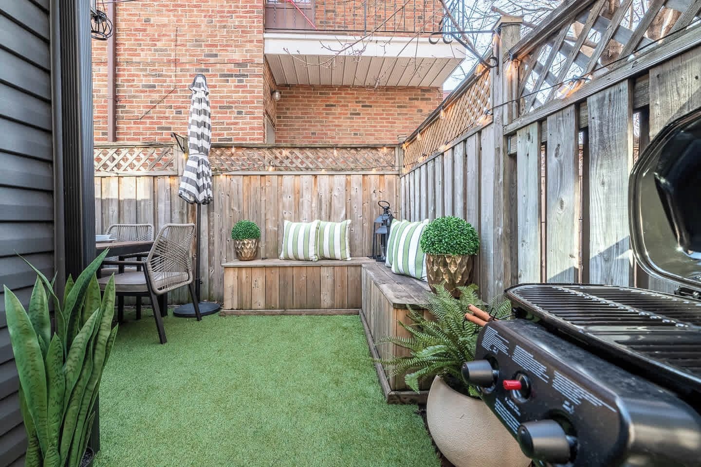 A backyard with a wooden deck, a barbecue grill, a seating area with cushions, and a small table.