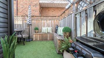 A backyard with a wooden deck, a barbecue grill, a seating area with cushions, and a small table.