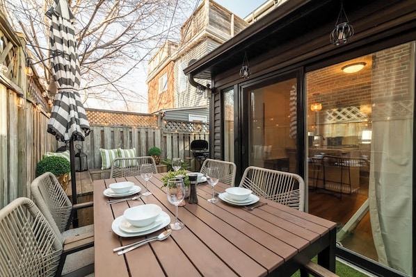 A wooden patio table set for dining with chairs, a grill, and a glass enclosure.