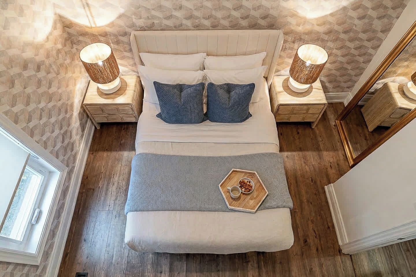A bed with a headboard, two bedside tables with lamps, a bedside tray with a cup and nuts, and a window.