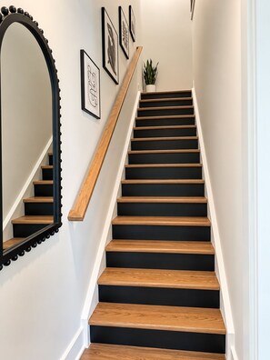 A staircase with wooden steps and black risers, a wooden handrail, and a wall-mounted mirror. - Modern Charlotte Retreat - Sleeps 10 - Near UNCC & Uptown (Charlotte)
