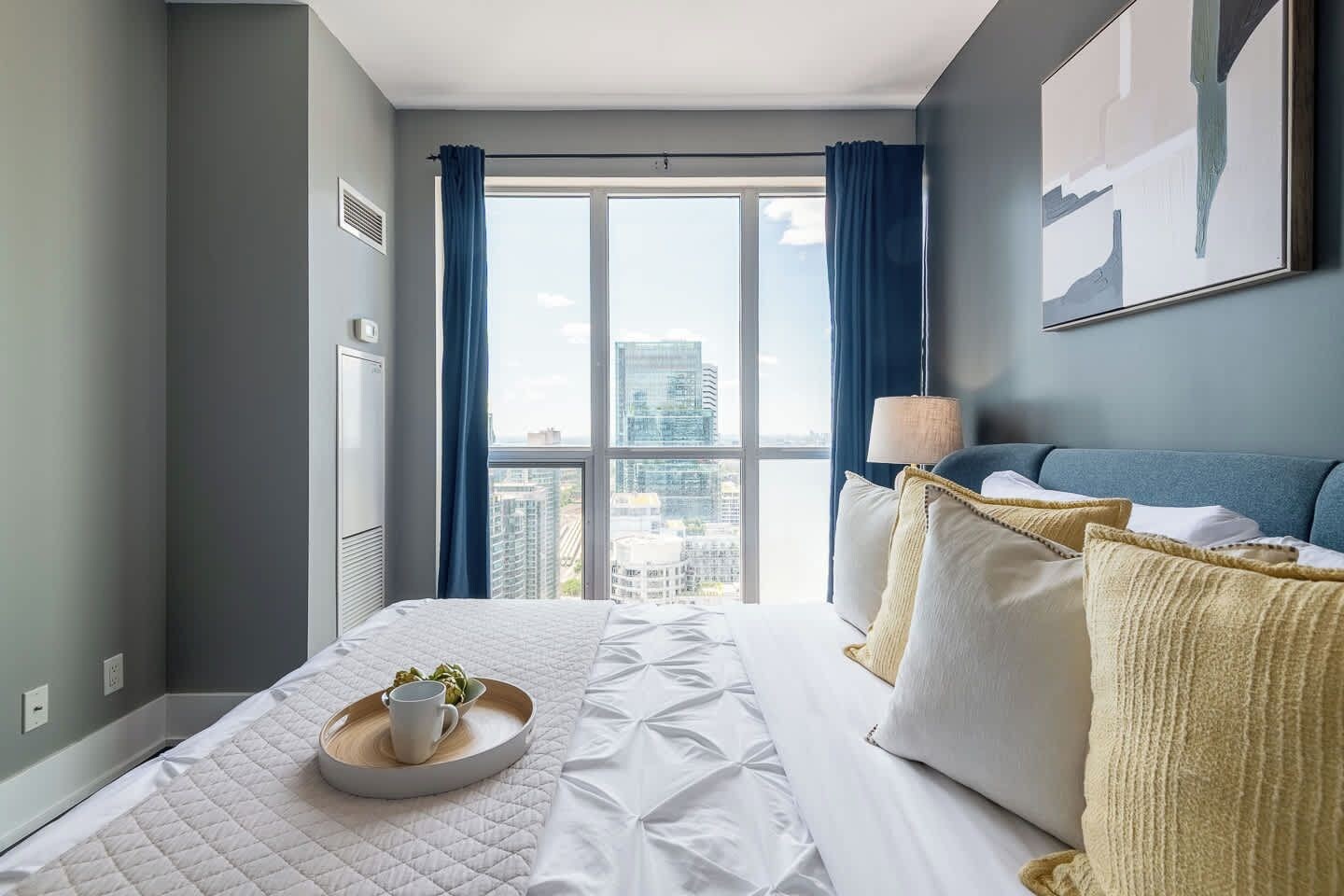 A hotel room with a large window, a bed with white bedding, a bedside table with a lamp, and a city view.