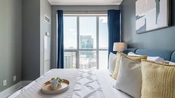 A hotel room with a large window, a bed with white bedding, a bedside table with a lamp, and a city view.
