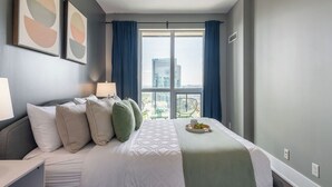 Habitación de hotel con cama, ventana con cortinas, vistas a la ciudad y una mesita con una bandeja de frutas.