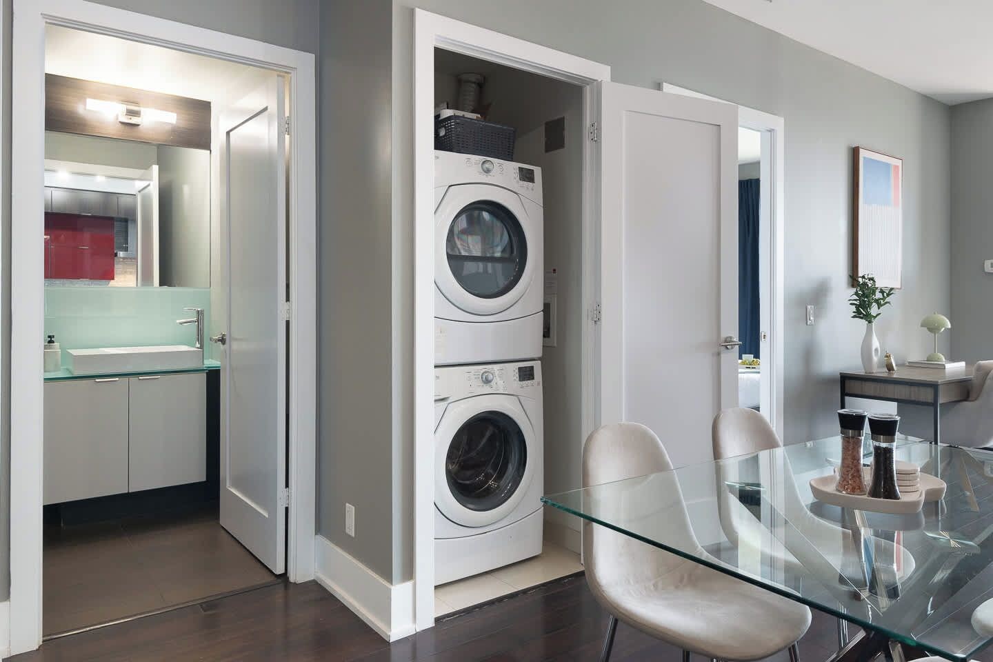 A modern laundry room with stacked washers and dryers, a glass dining table with chairs, and a kitchen visible through an open door.