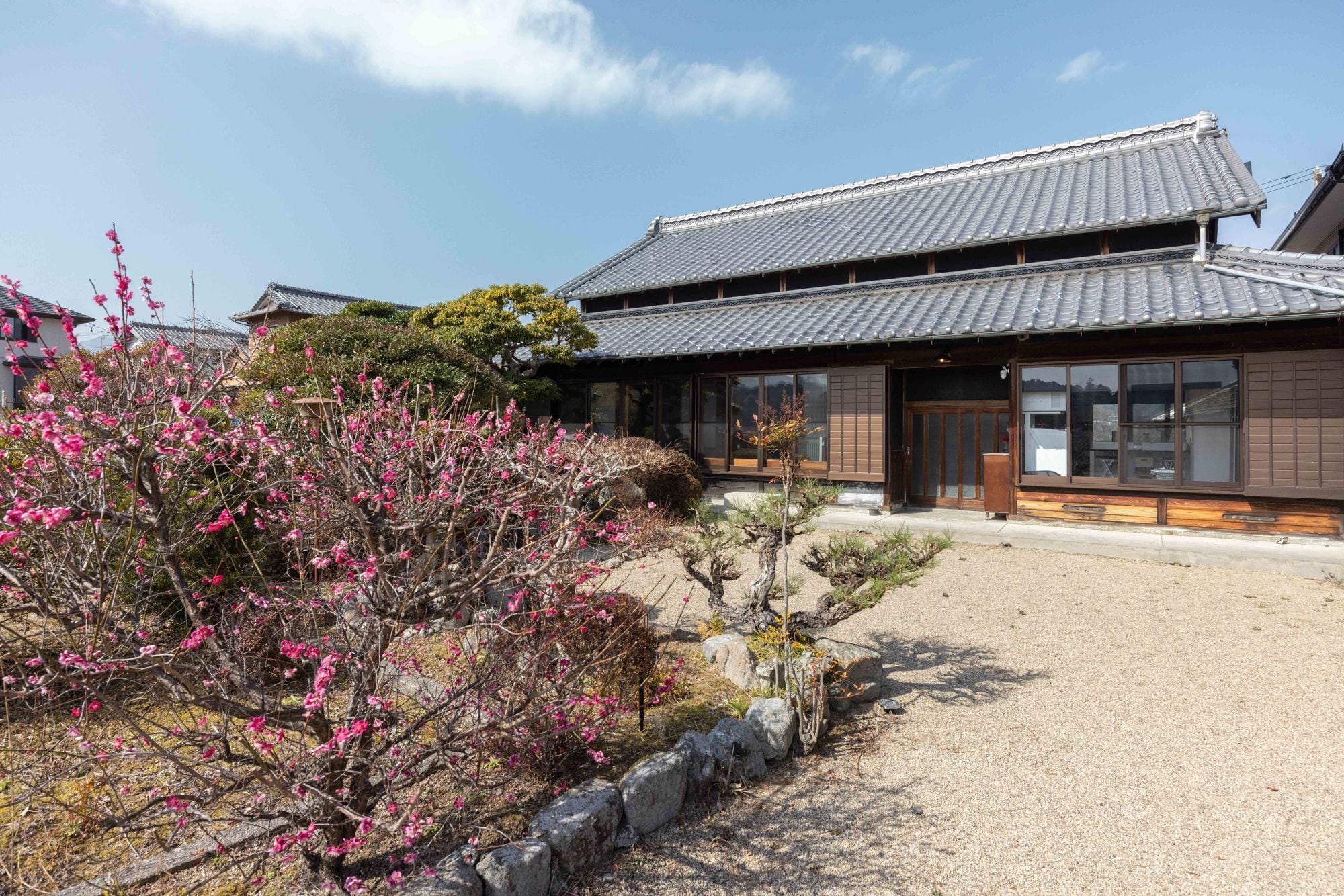 A traditional Japanese building with a tiled roof, a gravel driveway, and a garden with flowering trees.