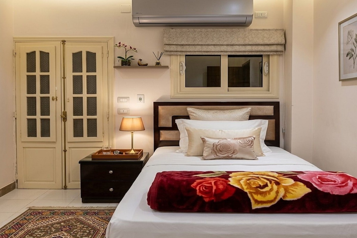 Et hotelværelse med en seng, et natbord, en lampe, et vindue med persienner, en fastgjort aircondition og en dør med glaspaneler.