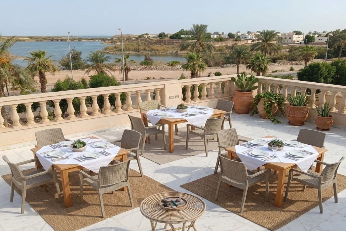 An outdoor dining area with tables and chairs, overlooking a beach and water.