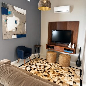 A modern living room with a flat-screen TV, a blue armchair, a large abstract painting, a woven rug, and a wicker ottoman.