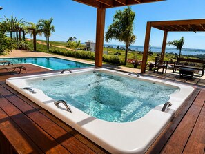 A hot tub with a wooden deck, overlooking a pool and a scenic view.