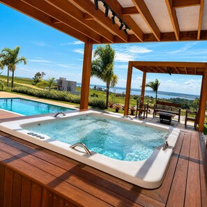 A hot tub with a wooden deck, overlooking a pool and a scenic view of a body of water and greenery.