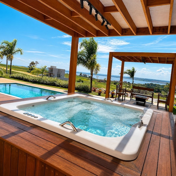 A hot tub with a wooden deck, overlooking a pool and a scenic view of a body of water and greenery.