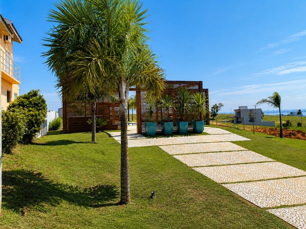 A modern outdoor area with a pathway leading to a wooden structure, surrounded by well-maintained grass and palm trees.