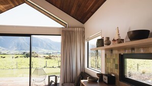 A room with a large sliding glass door leading to a grassy field, a wooden ceiling, a dining table with chairs, and a kitchen area with a tiled backsplash. - Infrared sauna, outdoor bath, close to town and mountain bike trails (Kaikōura)