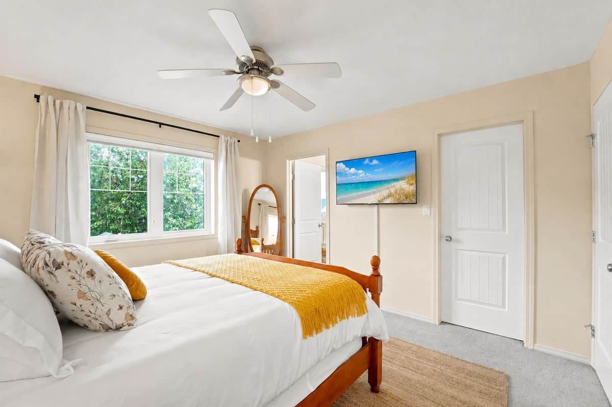 A bedroom with a bed, a ceiling fan, a window with curtains, a door, and a wall-mounted TV.
