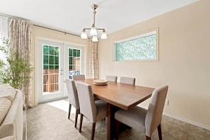 Un coin repas avec une table en bois, six chaises, un lustre, une plante en pot et une porte-fenêtre coulissante donnant sur un espace extérieur.