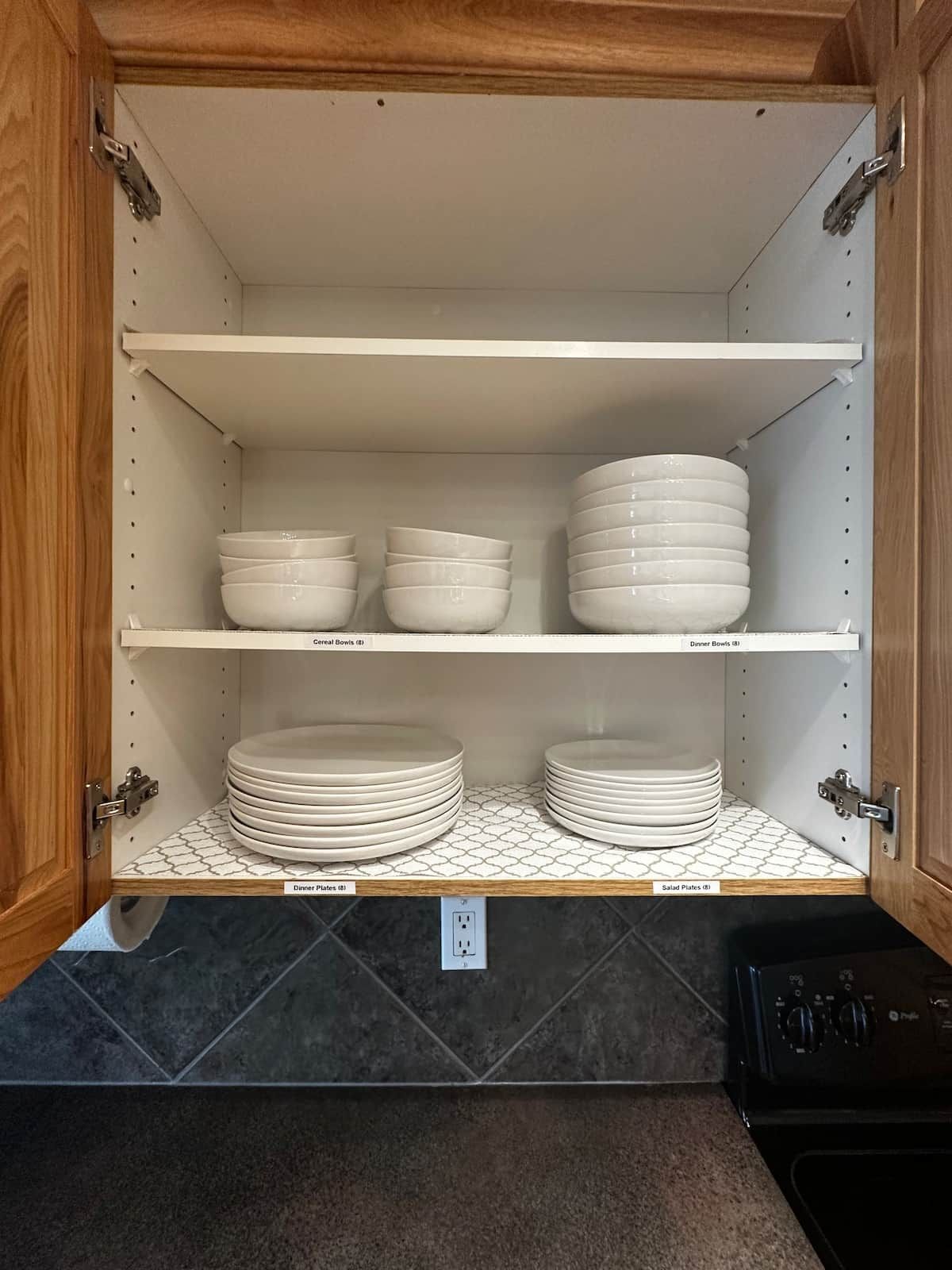 A kitchen cabinet with shelves holding various white bowls and plates, a tiled backsplash, and a black stove.