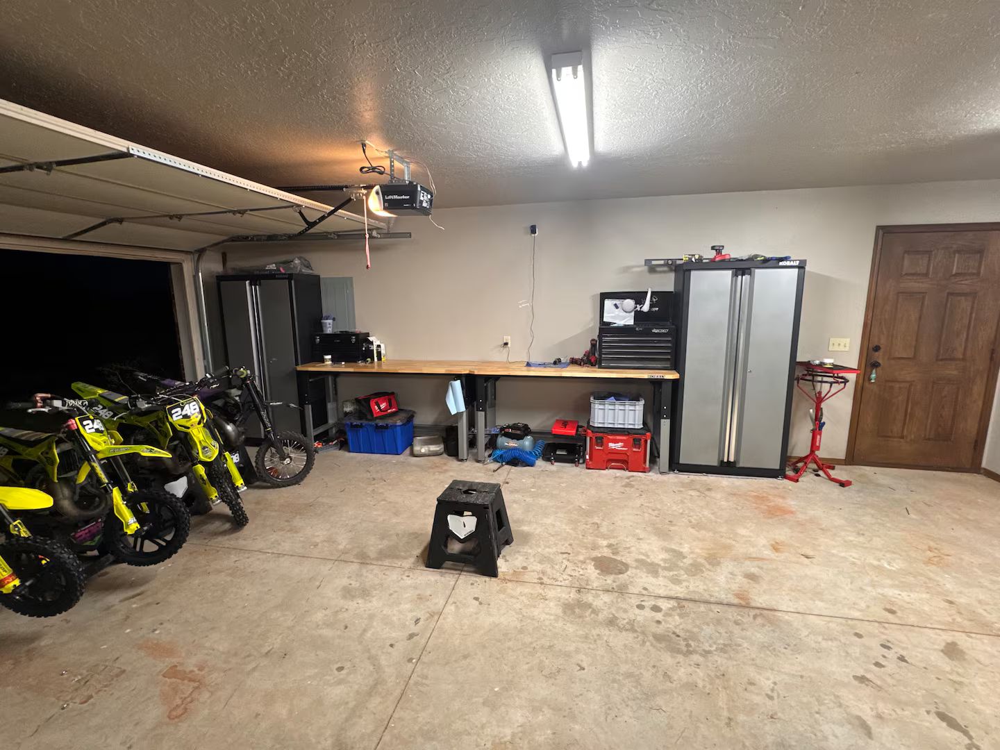 Un garaje con dos motocicletas, un banco de trabajo, un refrigerador y un proyector instalado en el techo.