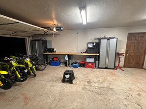 Un garaje con dos motocicletas, un banco de trabajo, un refrigerador y un proyector instalado en el techo.