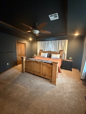 A bedroom with a wooden bed, a ceiling fan, a window with curtains, and a nightstand with a lamp. - HR Ranch... (Tecumseh)