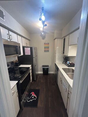 A compact kitchen with white cabinets, a microwave, a stove, and a refrigerator. There's a trash can and a coffee maker on the counter. - Relax & Recharge  in this Spacious Apartment / near the Medical Center & NRG! (Houston)