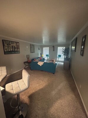 A living room with a blue sofa, a white chair, and a small table with a lamp. There is a painting on the wall and a window with blinds. - Relax & Recharge  in this Spacious Apartment / near the Medical Center & NRG! (Houston)