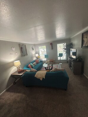 A cozy living room with a blue sofa, a small table with a lamp, and a window with blinds. - Relax & Recharge  in this Spacious Apartment / near the Medical Center & NRG! (Houston)