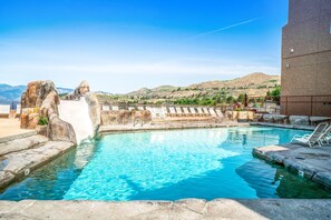 A pool area with clear water, a water slide, lounge chairs, and a view of a hilly landscape. - Grandview #615  Waterfront Chelan Condo (Chelan)