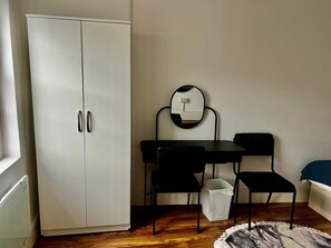 A room with a white wardrobe, a round mirror, a small table, two chairs, a waste bin, and a patterned rug. - Apartment Sleeps 2 with Wifi (London)