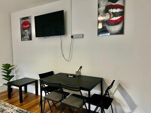 A modern living room with a flat-screen TV mounted on the wall, a dining table with four chairs, and two artwork pieces above the TV. - Apartment Sleeps 2 with Wifi (London)