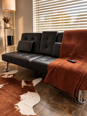 A modern living room with a black leather sofa, a brown knitted throw, and a remote control on a chair.